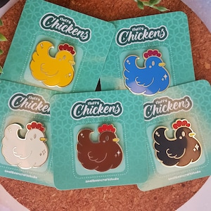 Op de afbeelding: Vijf emaille pins met een cartoon kippenontwerp in geel, blauw, wit, bruin en zwart. Elke pin heeft een rode kam en gouden accenten, weergegeven op turquoise kaarten met de tekst "Fluffy Chickens". De naam van de kunstenaar is ook zichtbaar.