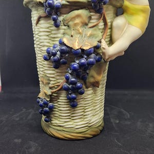 Puede incluir: Un jarrón de cerámica con un diseño de cesta tejida y una figura esculpida de un niño que alcanza un racimo de uvas azules. El jarrón está decorado con detalles dorados y tiene un asa.