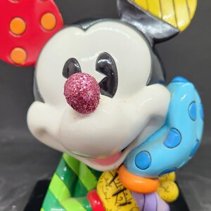K&ouml;nnte beinhalten: Eine farbenfrohe, handbemalte Keramikfigur von Mickey Mouse. Mickey tr&auml;gt ein gr&uuml;n-rot gestreiftes Hemd und einen blauen Handschuh mit wei&szlig;en Punkten. Seine Nase ist glitzernd pink. Die Figur steht auf einem schwarzen Sockel.