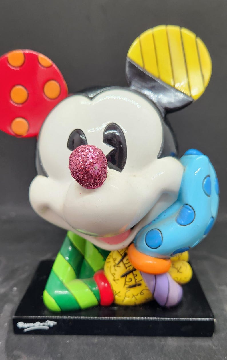 K&ouml;nnte beinhalten: Eine farbenfrohe, handbemalte Keramikfigur von Mickey Mouse. Mickey tr&auml;gt ein gr&uuml;n-rot gestreiftes Hemd und einen blauen Handschuh mit wei&szlig;en Punkten. Seine Nase ist glitzernd pink. Die Figur steht auf einem schwarzen Sockel.
