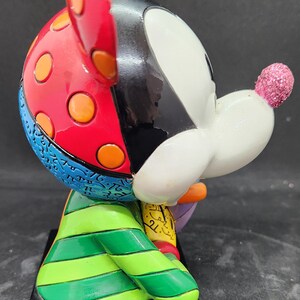 K&ouml;nnte beinhalten: Eine farbenfrohe, handbemalte Keramikfigur von Mickey Mouse. Die Figur sitzt mit einer Hand auf einem Buch. Mickeys Ohren sind rot mit orangefarbenen Punkten, sein K&ouml;rper ist gr&uuml;n-gelb gestreift und sein Gesicht ist wei&szlig; mit schwarzen Akzenten. Die Figur steht auf einem schwarzen Sockel.