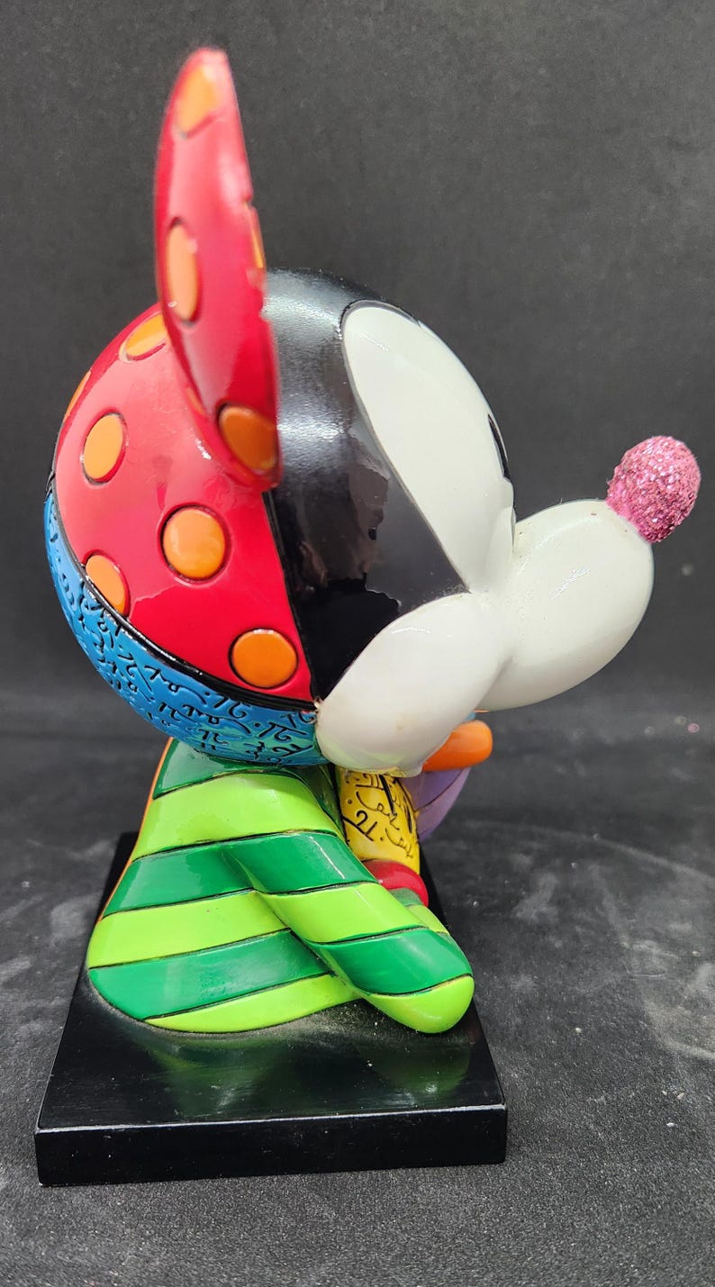 K&ouml;nnte beinhalten: Eine farbenfrohe, handbemalte Keramikfigur von Mickey Mouse. Die Figur sitzt mit einer Hand auf einem Buch. Mickeys Ohren sind rot mit orangefarbenen Punkten, sein K&ouml;rper ist gr&uuml;n-gelb gestreift und sein Gesicht ist wei&szlig; mit schwarzen Akzenten. Die Figur steht auf einem schwarzen Sockel.