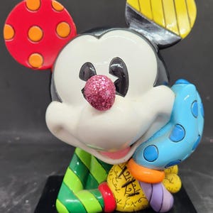 K&ouml;nnte beinhalten: Eine farbenfrohe Keramikfigur von Mickey Mouse, 16 cm hoch. Mickey tr&auml;gt ein gr&uuml;n-gelb gestreiftes Hemd und eine blau-wei&szlig; gepunktete M&uuml;tze. In seiner rechten Hand h&auml;lt er einen blau-wei&szlig; gepunkteten Handschuh.
