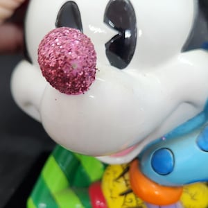 K&ouml;nnte beinhalten: Eine wei&szlig;e Keramikfigur von Mickey Mouse, der ein gr&uuml;n-rot gestreiftes Hemd, eine blaue Hose mit wei&szlig;en Punkten und eine glitzernde rosa Nase tr&auml;gt. Er h&auml;lt eine bunte Auswahl an Gegenst&auml;nden in seinen H&auml;nden.