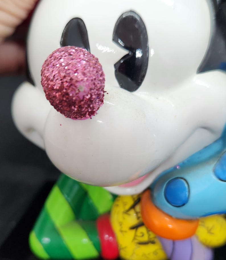 K&ouml;nnte beinhalten: Eine wei&szlig;e Keramikfigur von Mickey Mouse, der ein gr&uuml;n-rot gestreiftes Hemd, eine blaue Hose mit wei&szlig;en Punkten und eine glitzernde rosa Nase tr&auml;gt. Er h&auml;lt eine bunte Auswahl an Gegenst&auml;nden in seinen H&auml;nden.