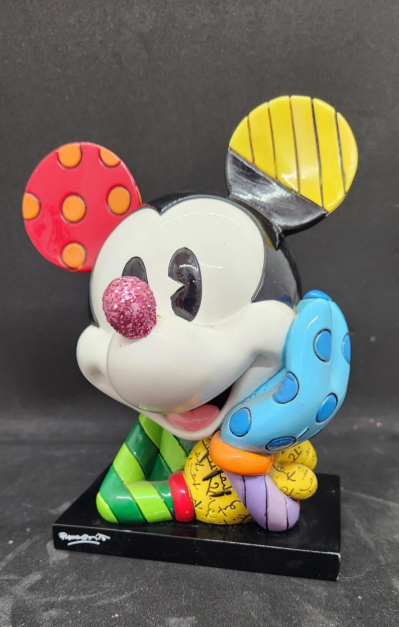 K&ouml;nnte beinhalten: Eine farbenfrohe, stilisierte Figur von Mickey Mouse mit einer pinkfarbenen Glitzernase. Er tr&auml;gt ein gr&uuml;n-rot gestreiftes Hemd und einen blauen Handschuh mit wei&szlig;en Punkten. Die Figur steht auf einem schwarzen Sockel mit der Unterschrift des K&uuml;nstlers.