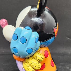 K&ouml;nnte beinhalten: Eine farbenfrohe Keramikfigur von Mickey Mouse mit schwarzem Kopf, wei&szlig;em Gesicht, blauem, gepunktetem Handschuh und orange-gelb gemusterter Kleidung. Die Figur steht auf einem schwarzen Sockel.
