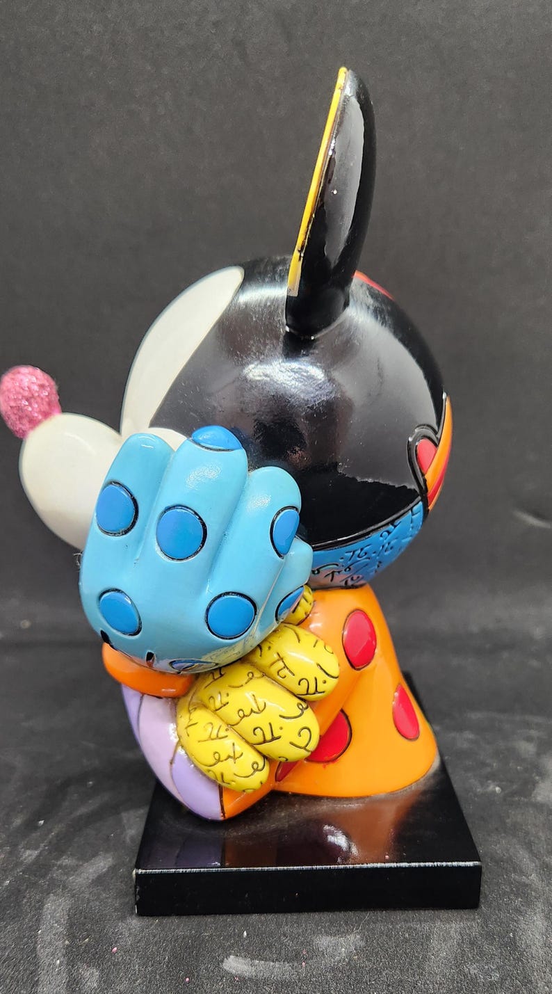 K&ouml;nnte beinhalten: Eine farbenfrohe Keramikfigur von Mickey Mouse mit schwarzem Kopf, wei&szlig;em Gesicht, blauem, gepunktetem Handschuh und orange-gelb gemusterter Kleidung. Die Figur steht auf einem schwarzen Sockel.