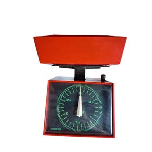 May include: A retro red and black kitchen scale with a circular dial displaying green numbers and markings. The scale has a rectangular base and a red bowl-shaped top. The dial reads in kilograms, with a maximum capacity of 3 kg.