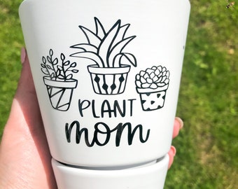 May include: White ceramic planter with a black design of three potted plants and the text "PLANT MOM".