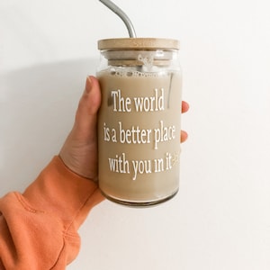 May include: A glass jar with a wooden lid and a metal straw filled with iced coffee. The jar has the text "The world is a better place with you in it" printed on it.