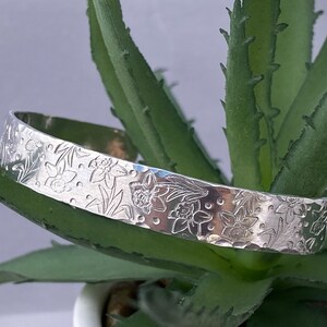 May include: A silver bangle bracelet with a hammered finish and an embossed design of daffodils.