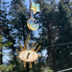 Può includere: Un acchiappasole con un cristallo bianco iridescente al centro, circondato da filo dorato. Sopra il cristallo, due cristalli iridescenti più piccoli, uno a forma di stella e l'altro a forma di mezzaluna.