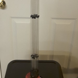 May include: A clear, vertical, cylindrical object with a red base and black connectors. The object is composed of multiple clear tubes held together by black connectors. The base is a bright red color. The object is standing on a black surface.