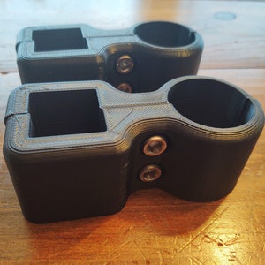 May include: Two black 3D printed clamps with a round opening and a square opening. The clamps have two silver screws on each side.