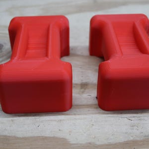 May include: Two bright red, rectangular objects with a T-shaped design. The objects have a textured surface and are placed side by side on a light-coloured wooden surface. The objects appear to be 3D-printed.