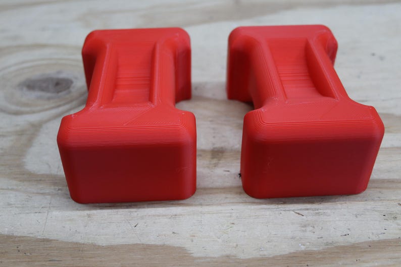 May include: Two bright red, rectangular objects with a T-shaped design. The objects have a textured surface and are placed side by side on a light-coloured wooden surface. The objects appear to be 3D-printed.