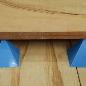 May include: Two bright blue, triangular supports holding up a piece of wood. The supports are evenly spaced and appear to be 3D-printed. The wood is a light brown color with a darker brown edge.