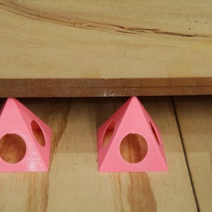 May include: Two pink, pyramid-shaped woodworking supports with circular cutouts are positioned under a wooden board. The supports are designed to elevate the board, providing space for various woodworking tasks. The supports are made of a solid material.