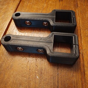 May include: Two black 3D printed plastic brackets with screw holes and a square opening. The brackets are on a wooden surface.