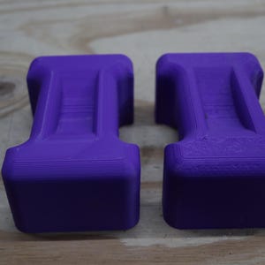 May include: Two purple, rectangular objects with a unique shape, possibly weights or supports. The objects have a solid, blocky base and a central, raised section. The colour is a vibrant, saturated purple, and the objects are placed on a wooden surface.