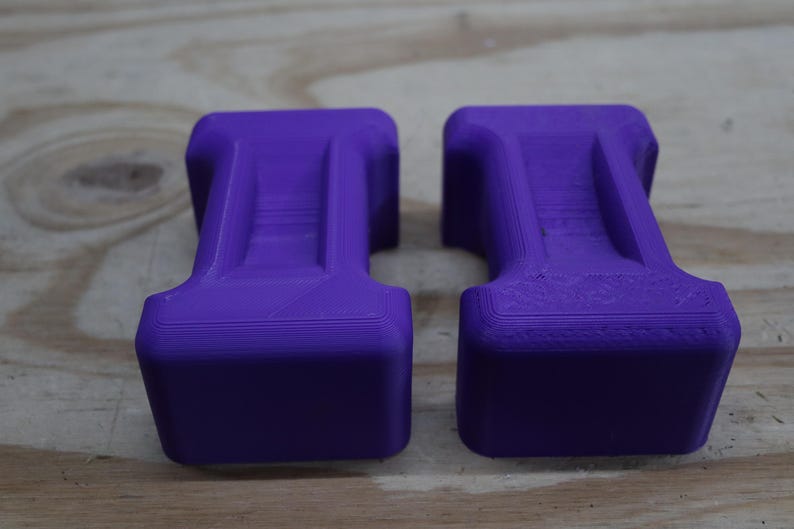 May include: Two purple, rectangular objects with a unique shape, possibly weights or supports. The objects have a solid, blocky base and a central, raised section. The colour is a vibrant, saturated purple, and the objects are placed on a wooden surface.