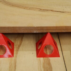 May include: Two bright red, pyramid-shaped woodworking supports with circular cutouts are positioned under a wooden board. The supports are designed to elevate the board, providing space for various woodworking tasks.