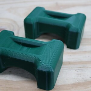May include: Two dark green, rectangular objects with a handle-like indentation on top. The objects are 3D-printed and have a textured surface. They are placed on a light brown wooden surface.