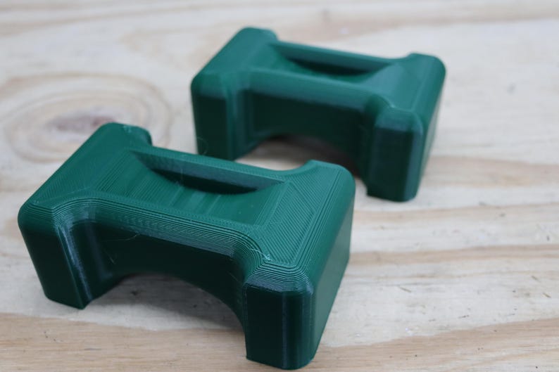 May include: Two dark green, rectangular objects with a handle-like indentation on top. The objects are 3D-printed and have a textured surface. They are placed on a light brown wooden surface.