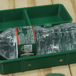 May include: A clear plastic water bottle inside a dark green, rectangular container with a hinged lid. The bottle has a red and green label. The container is open, revealing its interior.