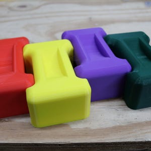 May include: Four brightly coloured, geometric objects in red, yellow, purple, and green. Each has a rectangular base and a curved top with a groove. They are arranged on a light wood surface.