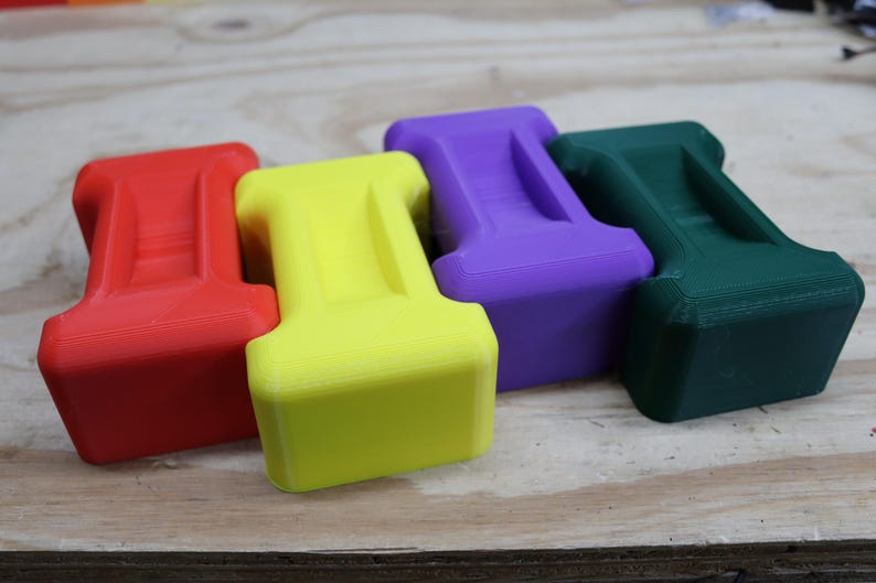 May include: Four brightly coloured, geometric objects in red, yellow, purple, and green. Each has a rectangular base and a curved top with a groove. They are arranged on a light wood surface.
