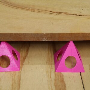 May include: Two bright pink, 3D-printed pyramid-shaped supports with circular cutouts. They are holding up a piece of wood. The supports are on a light-colored wooden surface.