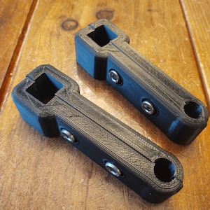 May include: Two black 3D printed handles with a square opening and a round opening at the end. Each handle has three silver screws.