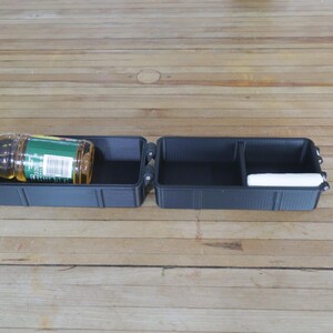 May include: A black, divided storage container with a hinged lid, holding a bottle of liquid and a white item. The bottle has a yellow cap and a green label. The container is open and sits on a wooden surface.