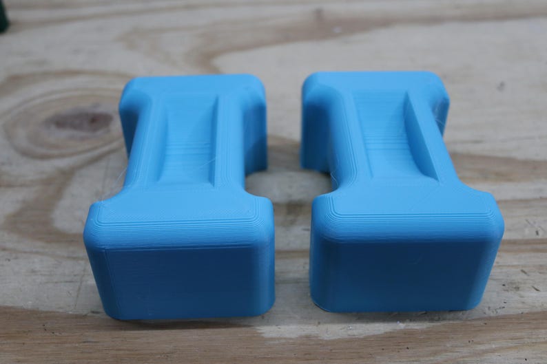 May include: Two light blue, 3D-printed objects with a unique, stylized shape. Each has a rectangular base and a central, raised section. The objects are displayed on a light brown wooden surface.