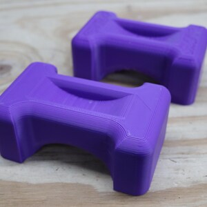 May include: Two purple, rectangular objects with a curved handle-like indentation on top. The objects are made of a solid material and have a textured surface. They are resting on a light-coloured wooden surface.