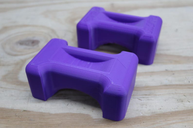 May include: Two purple, rectangular objects with a curved handle-like indentation on top. The objects are made of a solid material and have a textured surface. They are resting on a light-coloured wooden surface.