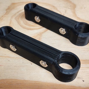 May include: Two black 3D printed metal brackets with silver screws. The brackets are designed to be attached to a surface and have a round opening on one end.