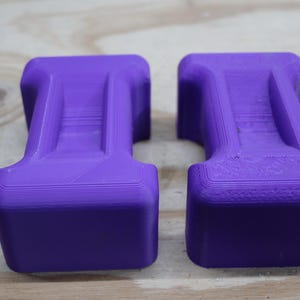 May include: Two identical, solid purple objects with a rectangular base and a curved top. The objects have a layered appearance and are set against a light wood grain background. The objects appear to be 3D printed.
