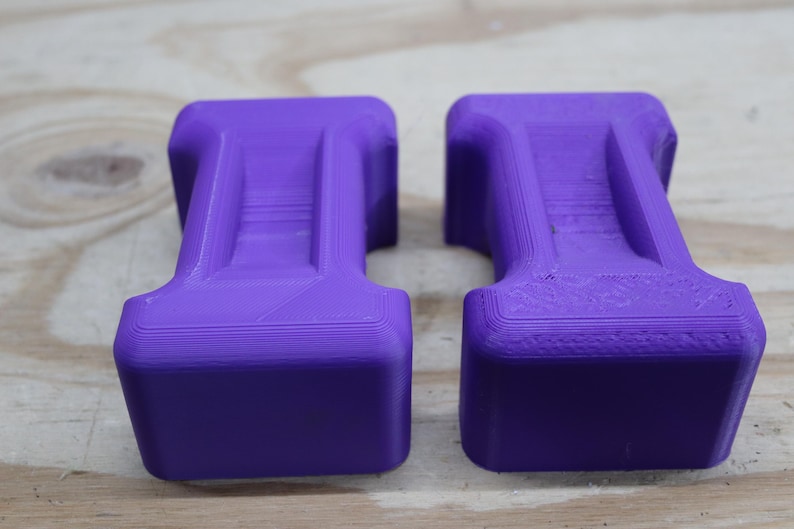 May include: Two identical, solid purple objects with a rectangular base and a curved top. The objects have a layered appearance and are set against a light wood grain background. The objects appear to be 3D printed.
