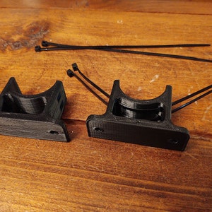 May include: Two black 3D printed wall mounts with a circular opening. The mounts are designed to hold pipes or other cylindrical objects.