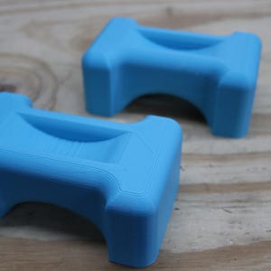 May include: Two light blue, 3D-printed objects. Each has a rectangular top with a curved indentation and a rounded arch underneath. The objects are made of plastic and sit on a wooden surface.