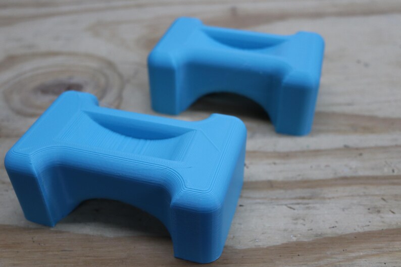 May include: Two light blue, 3D-printed objects. Each has a rectangular top with a curved indentation and a rounded arch underneath. The objects are made of plastic and sit on a wooden surface.