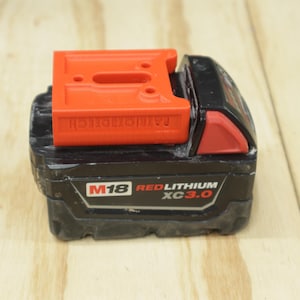 May include: A black and red M18 RedLithium XC3.0 battery with an orange adapter on top. The adapter has the text "RADIO-TECH" printed on it. The battery is on a light wood surface.