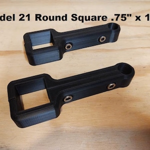 May include: Two black plastic 3D printed square-shaped objects with rounded edges and a hole at one end. The objects are 1.9 cm by 3.6 cm. "Model 21 Round Square .75" x 1.41" is written in white text at the top of the image.