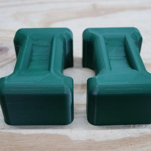 May include: Two dark green, rectangular objects with a unique, stepped design. The objects have a textured surface and are placed side-by-side. The objects are likely 3D-printed and sit on a light-coloured wooden surface. The objects have a handle-like shape.