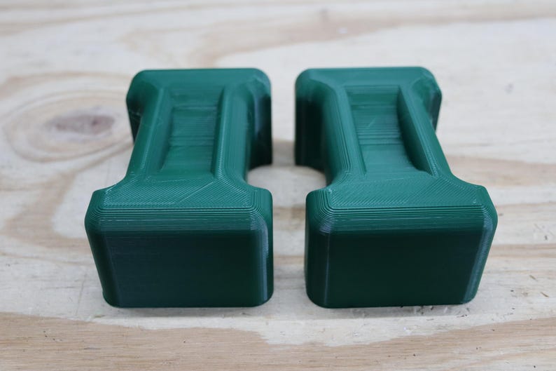 May include: Two dark green, rectangular objects with a unique, stepped design. The objects have a textured surface and are placed side-by-side. The objects are likely 3D-printed and sit on a light-coloured wooden surface. The objects have a handle-like shape.
