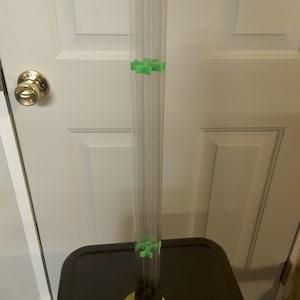 May include: A clear plastic tube with green plastic connectors at the top and bottom. The tube is sitting on a black surface with a yellow base.