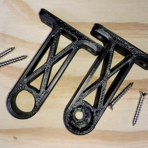 May include: Two black, 3D-printed brackets with a geometric design, each featuring a circular hole and a mounting surface. Several silver screws are scattered around the brackets on a light wood surface.